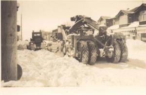 Crawler tractor and Scraper clearing snow on 12th ave. across from the Campbell Block, and Harold J. Clasky's store, on end of building on right, and H. McLeod Company, Insurance & Financial Agents on right side. This building sat where B of M sits today, 2017, south of the Smith Block. 